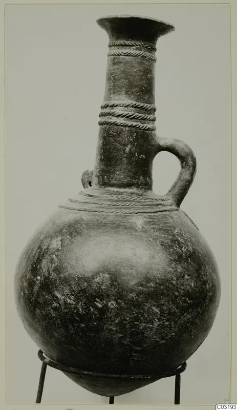 Globular jug with a tall neck, flaring rim, twisted raised bands and a single loop handle, standing on a metal stand.