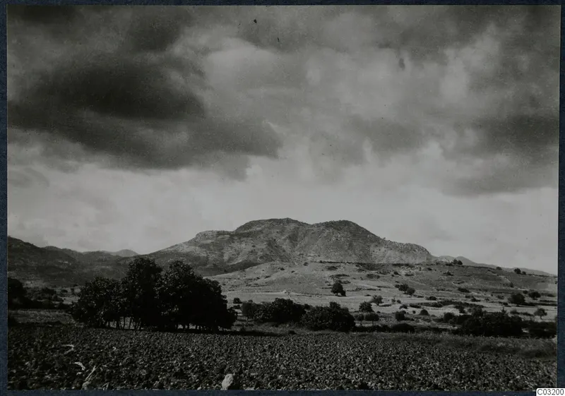 Dark, cloud-filled sky over a rocky hill with a dark tree cluster and a cultivated field in the foreground, at Soli.