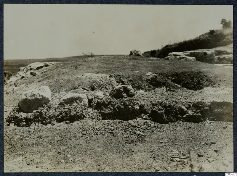 Theatre at Soli: stony ground with low earth mounds and rubble in the foreground, a scrubby hillside with a tree at right.