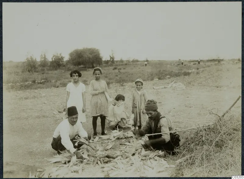 Two men shucking maize cobs on dry ground at Soli, with four children standing and crouching behind them.