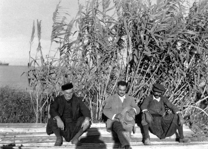 Three men crouching side by side on a wooden plank surface in front of tall reeds, with the sea on the left, at Karavostassi, Soli.