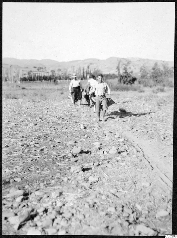 Three men walk across a stony, dry field at Soli, with hills and trees in the background.