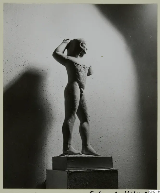 Limestone statue of a standing nude male figure with one arm raised to the head, photographed on a wooden pedestal