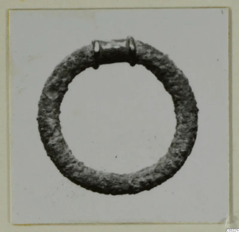 Corroded metal ring with a collar-like band and small loops at the top, photographed on a pale background.