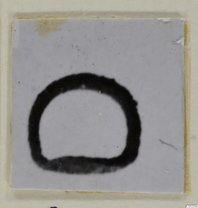 Small square of pale card or ceramic with a thick black ring-shaped mark, open at the bottom, on a cream backing
