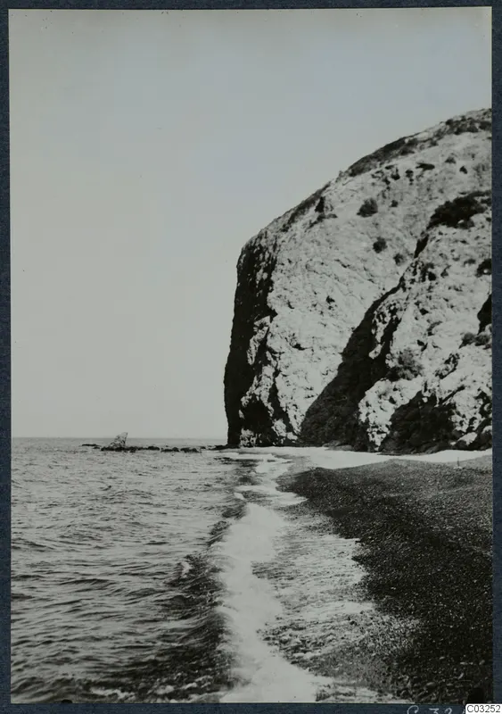 Steep pale rock cliff rising above a pebbly shore, with waves running in from the sea on the left.