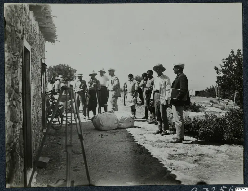 Men standing on a dirt track beside a stone building, with a rolled bundle on the ground and a bicycle against the wall at Vouni.