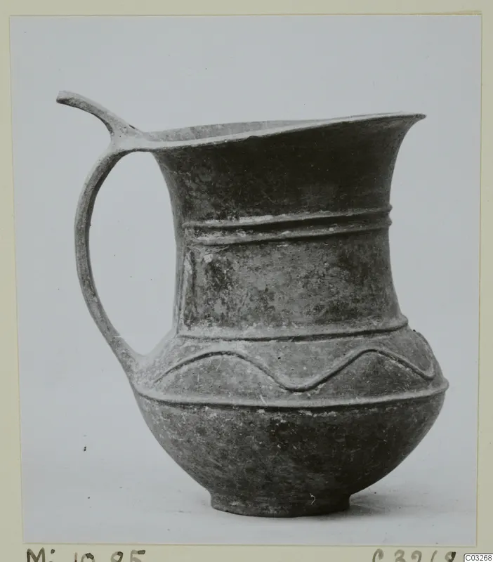 Pottery jug with a single handle, a pinched spout and raised horizontal bands, photographed on a plain background.