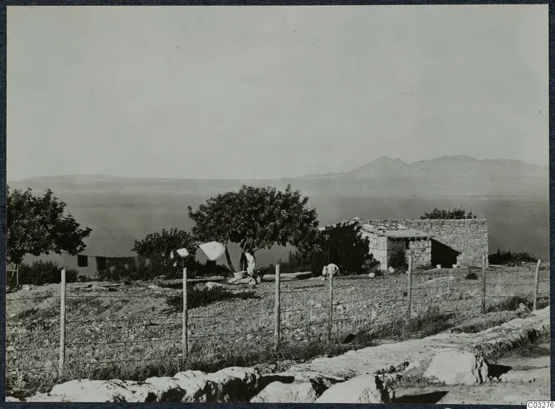 Low stone building with a flat roof beside a tree, with two people near them at Vouni, sea and hills behind