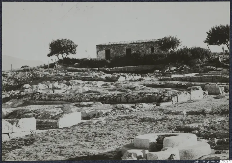 Ruined stone walls and foundations at Vouni Palace, with a small stone building and trees behind.
