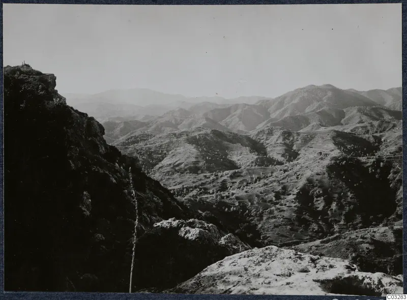 Mountain landscape seen from a rocky height at Vouni, with hazy sky, a dark cliff edge and scrub-covered slopes.