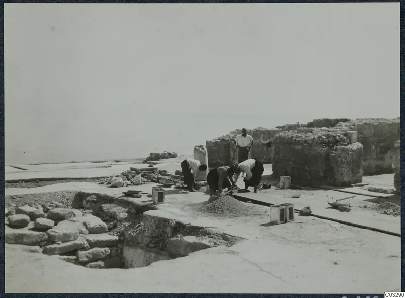 Workers repairing stonework in Room 47 at Vouni, with a standing man watching and the sea beyond the ruins.