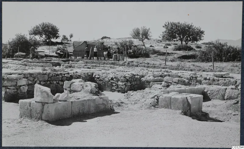 Excavated stone walls at the Vouni palace, with a canvas tent, three people and a wire fence behind