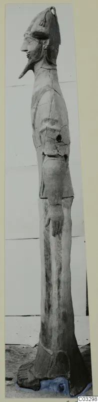 Tall terracotta standing figure in profile, with a conical headdress, long pointed nose and pointed beard, shown against a pale wall.