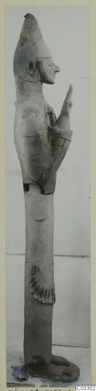 Tall terracotta figure of a standing figure in profile facing right, one arm raised, with a conical head covering.