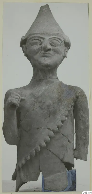 Terracotta figure with a tall pointed cap, large round eyes and a fringed diagonal garment across the chest