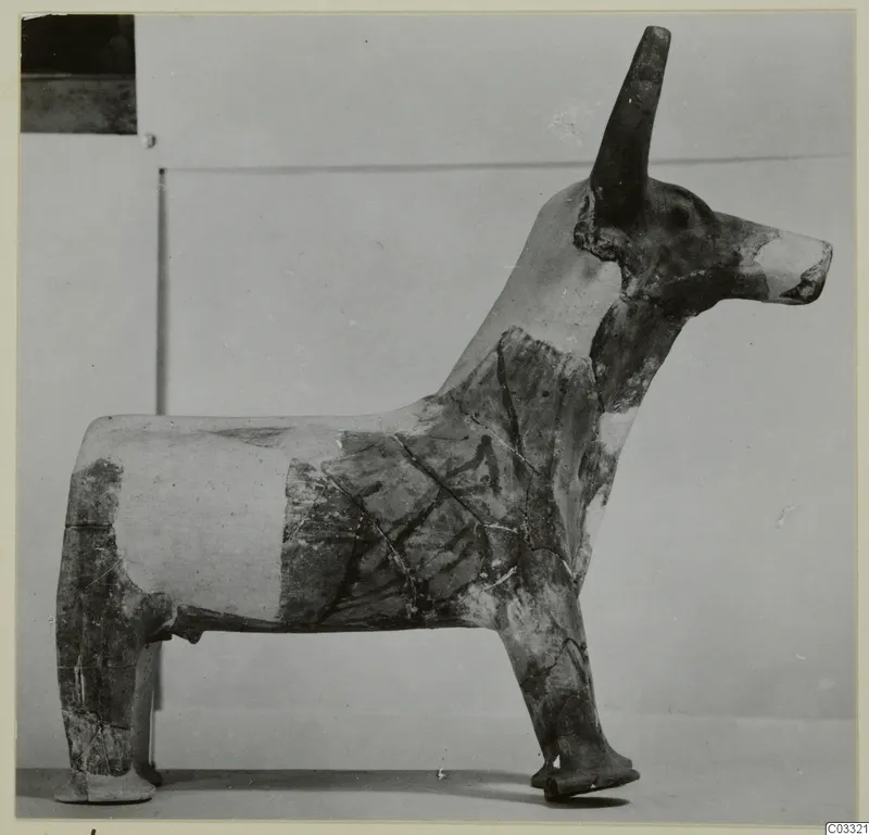 Terracotta figurine of a horned animal standing on four legs, with dark painted bands on a pale surface