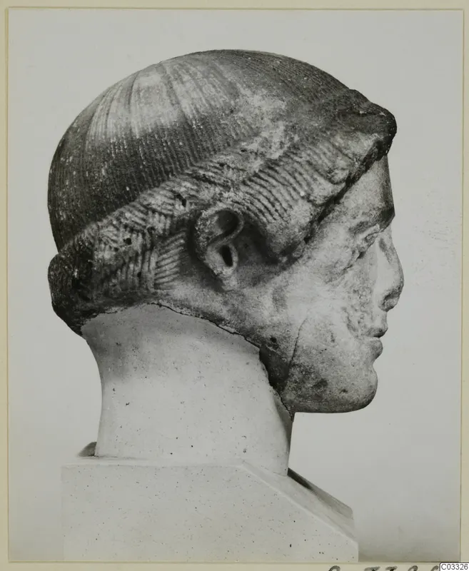 Carved stone head in profile, with a band of incised hair over the forehead and ear, on a plain pedestal.