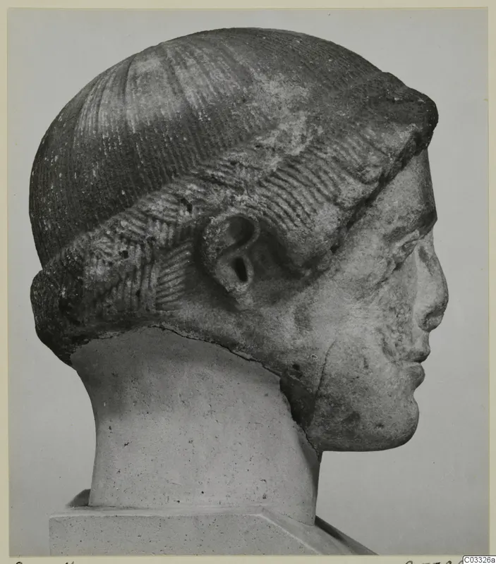 Carved head in profile facing right, with a tall rounded headdress of incised lines and a modelled ear.