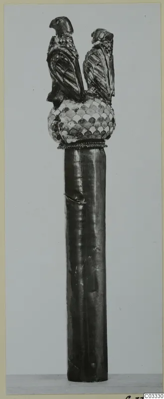 Upright ornamented object: a dark cylindrical shaft topped by a scale-patterned knob and two bird figures