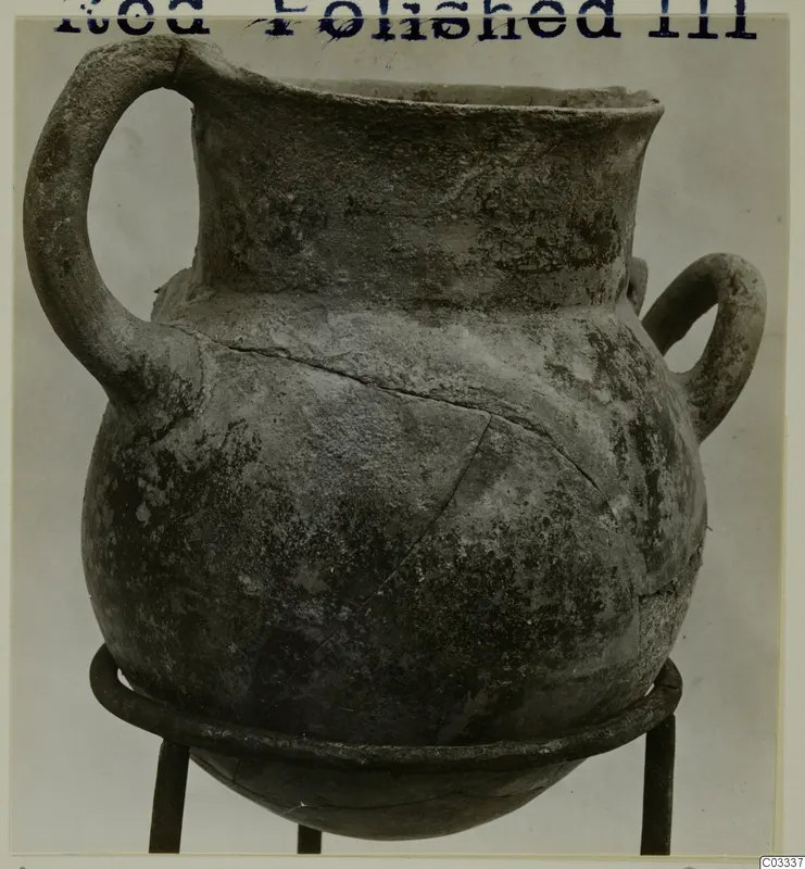 Red Polished III jug with a high loop handle and a second small handle, standing on a metal stand.