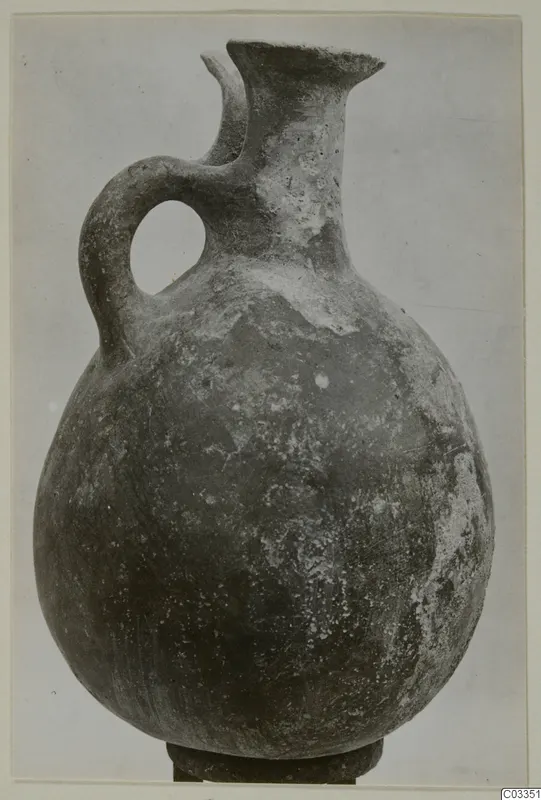 Pottery Red Polished III jug with a rounded body, tall neck, lipped spout and strap handle, standing on a dark base