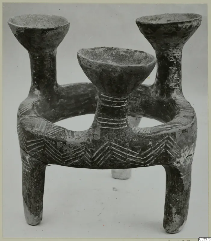 Dark pottery vessel with a ring-shaped body, three upright open cups and three legs, incised with chevron and band patterns