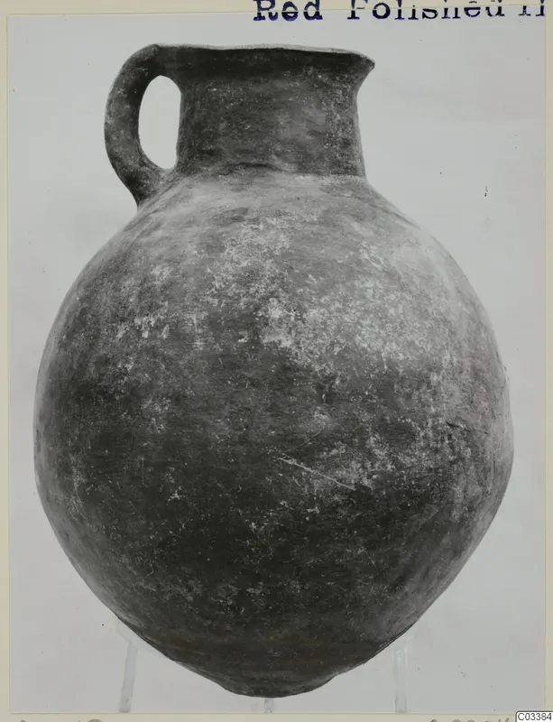 Red Polished II jug with a single loop handle, a short flaring neck and a rounded body, photographed on a clear stand
