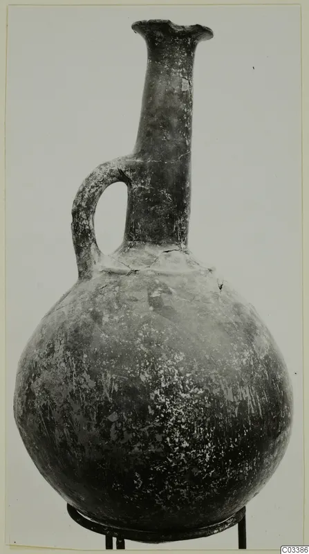 Pottery jug with a tall neck, flared rim, single loop handle and round body, standing on a metal stand.