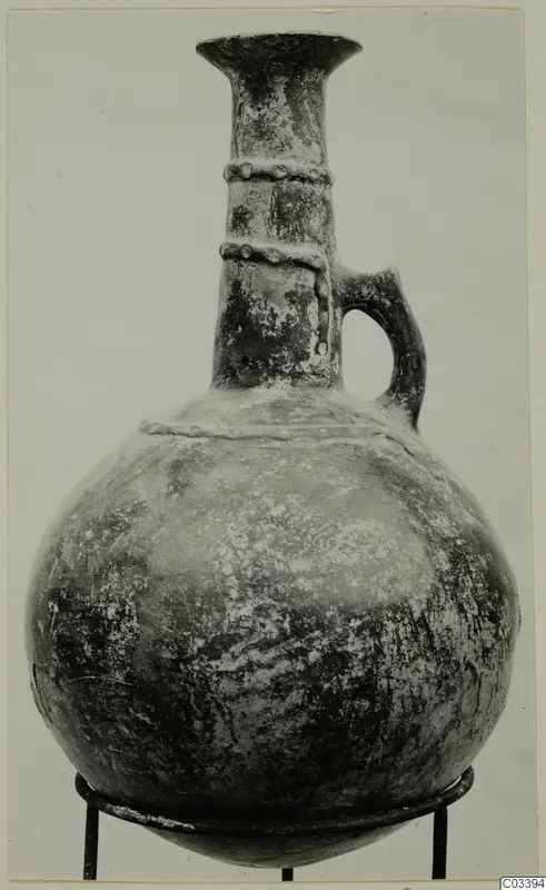 Red Polished II jug with a tall flared neck, two applied ridges on the neck, and a single loop handle, shown on a metal stand.