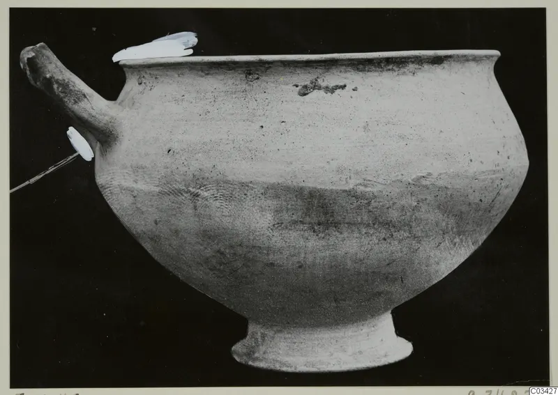 Hand-made pottery bowl with a horizontal spout-like handle on a short ring foot, photographed against a black background.