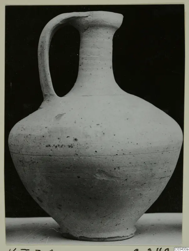 Pale ceramic jar with a single loop handle and tall neck, photographed against a black background