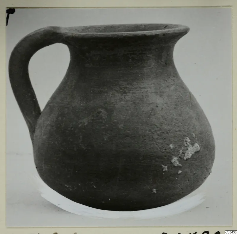Dark grey pottery jug with a single loop handle and wide rim, photographed against a plain background.