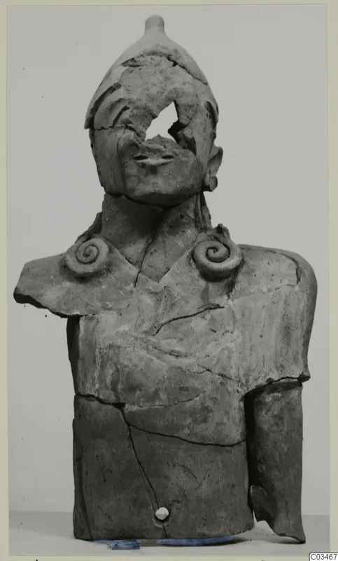Terracotta bust of a figure with a rounded headdress, spiral shoulder ornaments and a broken face, against a plain grey background