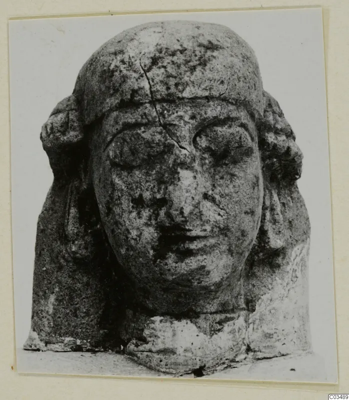 Limestone sculpted head with a flat headdress, heavy-lidded eyes and side locks, photographed on a card mount