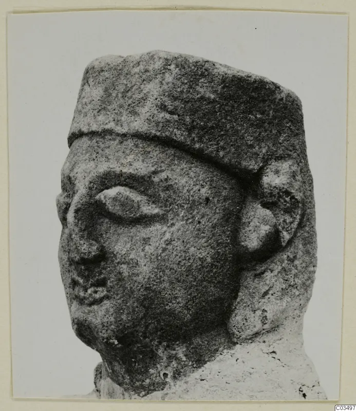 Limestone sculpture of a head wearing a flat-topped cap, with a large almond-shaped eye and a rounded ear