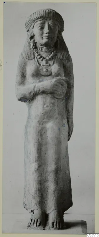 Standing female terracotta figure with arms folded across her chest, wearing a bead necklace and pendant.
