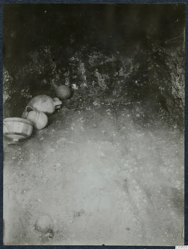 Pottery vessels lying on the sandy floor of an excavation at Stylli, against a rough rock wall.