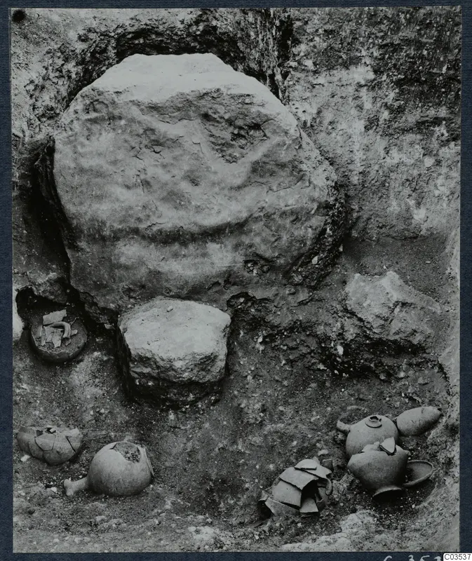 Excavation at Stylli with several broken pots and objects lying on the excavated floor beside a large stone