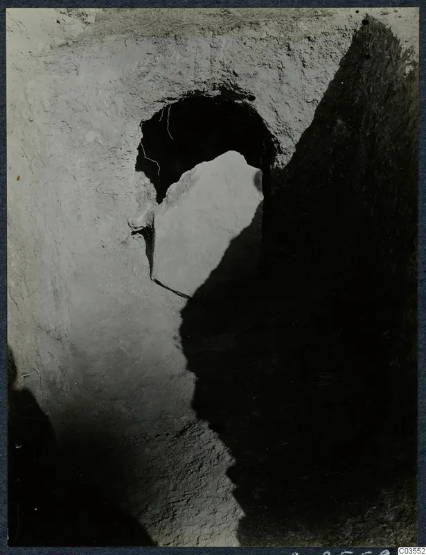 Excavation at Stylli with a large stone slab leaning in front of a dark opening cut into earth