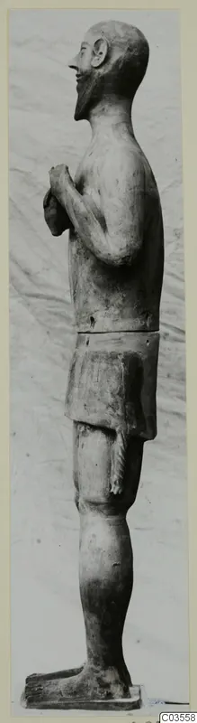 Side view of a standing bearded terracotta statue with hands held at the chest