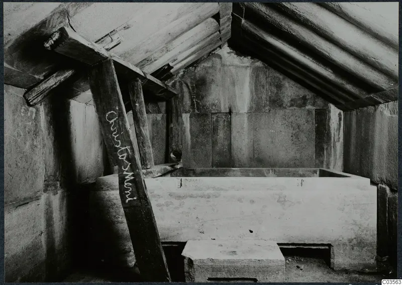 Built tomb chamber with timber props under a sloping roof and a large rectangular stone block in the centre.
