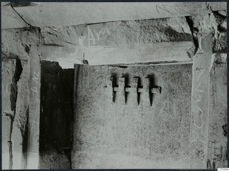 Interior of a built tomb chamber with scratched graffiti on stone slabs, cracks, and a dark opening to one side.