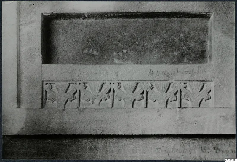 Close view of carved stone with a row of five fan-shaped reliefs below a recessed panel and faint lettering.