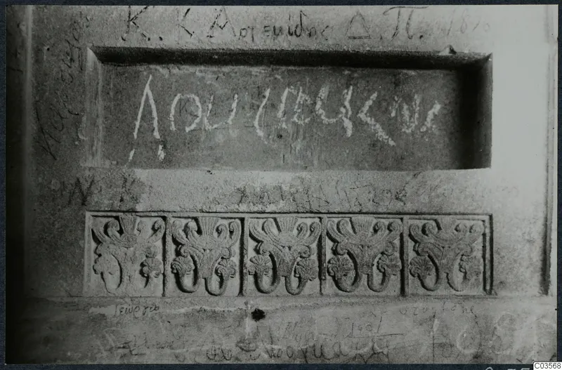 Carved stone wall in a built tomb chamber, with a recessed inscribed panel, five relief ornaments below and scratched graffiti