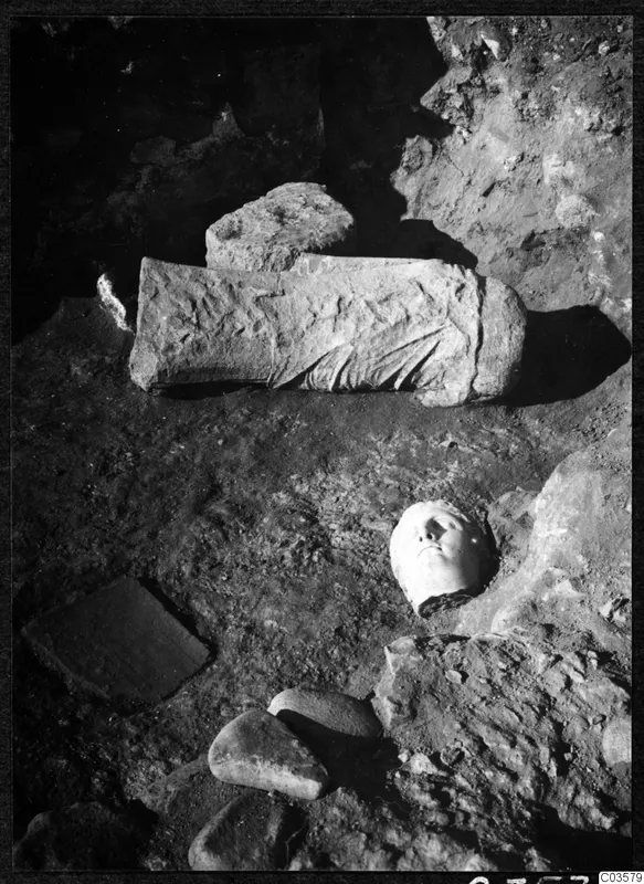 Carved stone head and a long carved stone fragment lying among rubble in excavated ground at Cholades, Soli