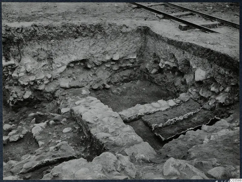 Excavation at Soli showing a stone wall and paved stone features in a trench, with a railway line at the top edge