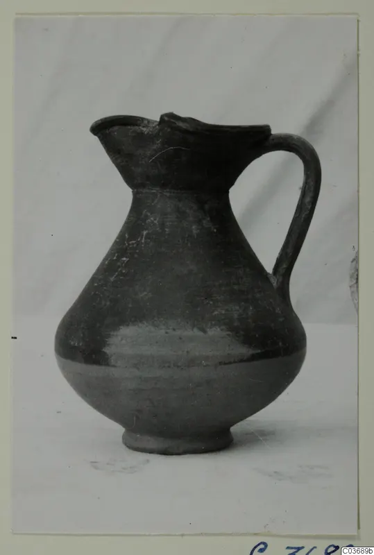 Jug with a single handle and a trefoil-like pinched spout, dark surface, photographed on a plain background.