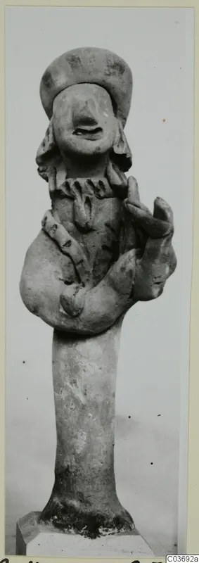 Terracotta figurine of a standing figure with bent arms and a wide cap-like headdress, set on a plinth