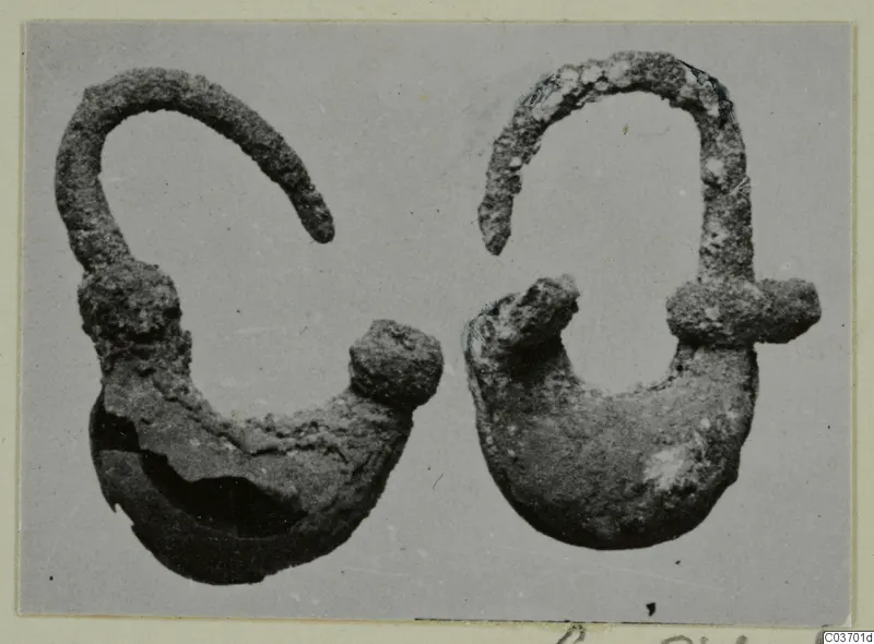 Two corroded metal objects from Vouni, each with an arched wire arm and a rounded lower body, shown on a grey ground.
